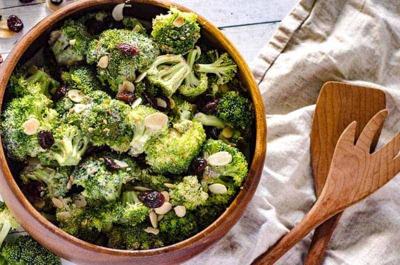 overhead view of wooden salad bowl full of raw broccoli salad with wooden salad utensils next to it