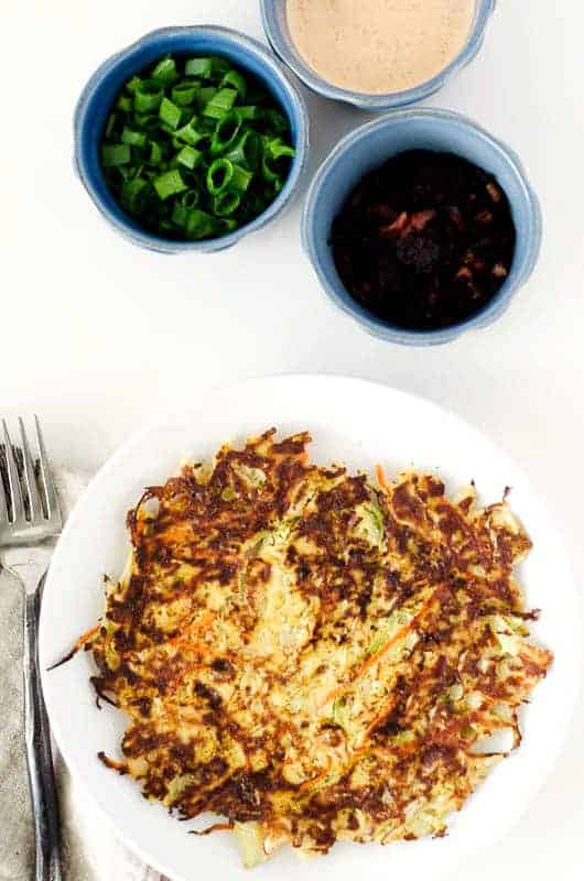 Overhead view of Okonomiyaki (Japanese Cabbage Pancake) on white plate with toppings in blue bowls nearby