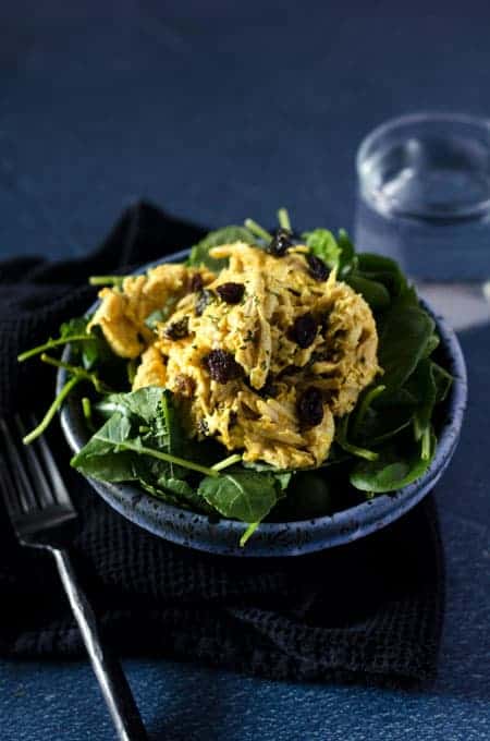 side view of a black and blue bowl full of greens topped with curry cashew whole30 chicken salad with a glass of water in the background