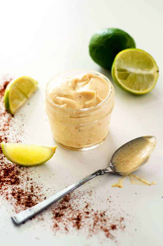 Jar of chipotle lime mayo surrounded by sliced limes, chili powder, and a spoon covered in mayo