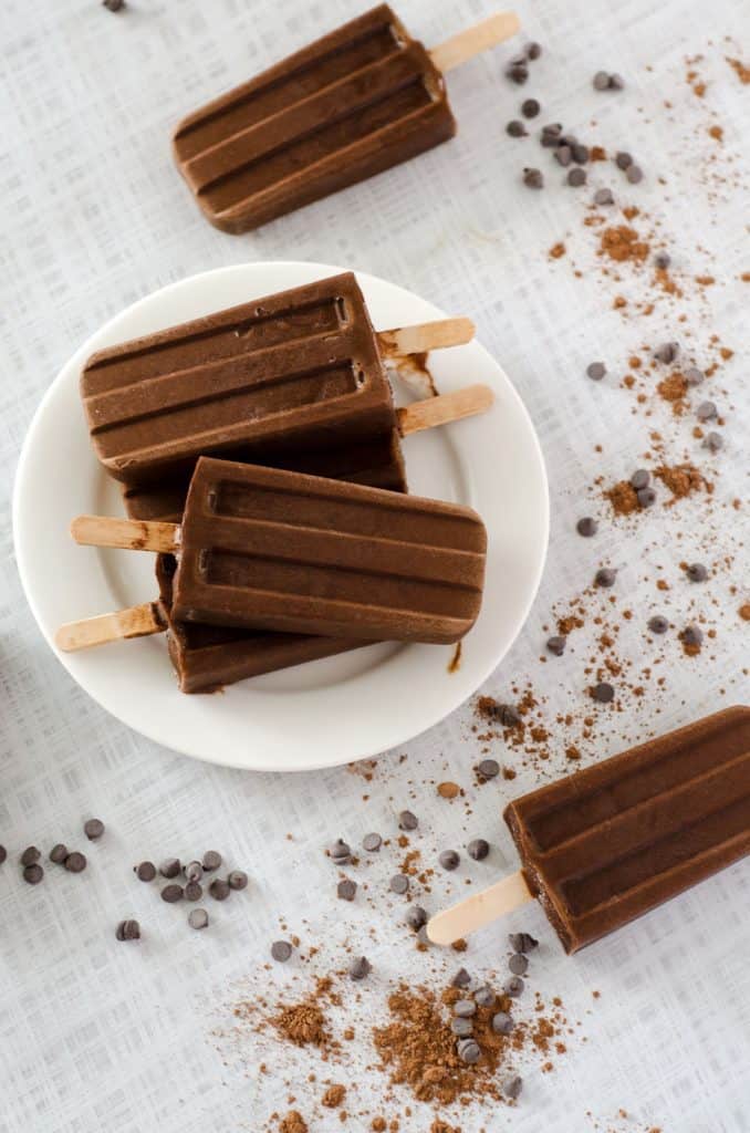 Overhead view of chocolate keto popsicles stacked on a white plate on a tabletop surrounded by cocoa powder and chocolate chips