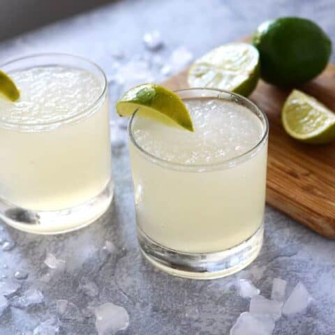 two glasses of frozen limeade surrounded by ice and a cutting board with limes