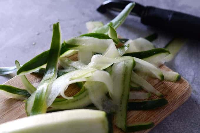 Cucumber ribbons on cutting board with vegetable peeler in the background