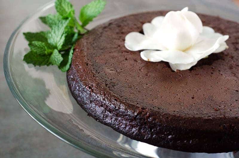 side view of paleo flourless chocolate cake on a glass cake stand topped with a white flower