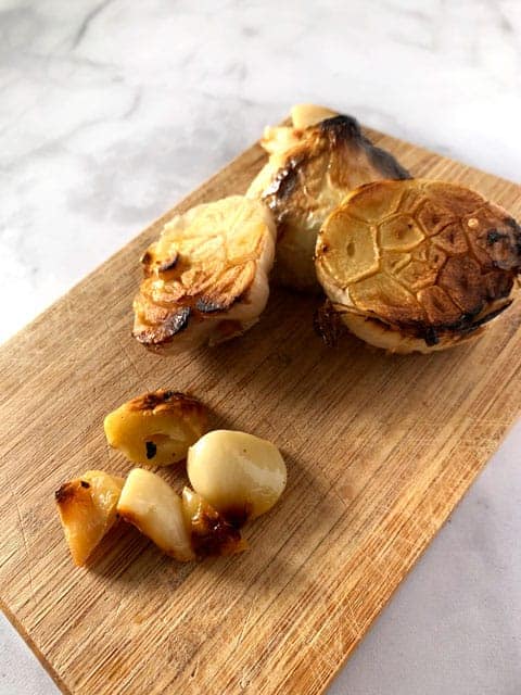 Side view of wooden cutting board with heads and cloves of roasted garlic