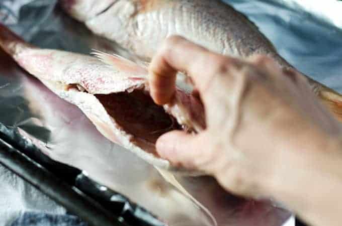 raw whole fish on a pan lined with foil with a hand holding the fish open and showing the empty body cavity