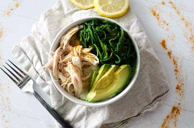 overhead view of a bowl full of chicken, greens, and avocado surrounded by turmeric and lemon slices