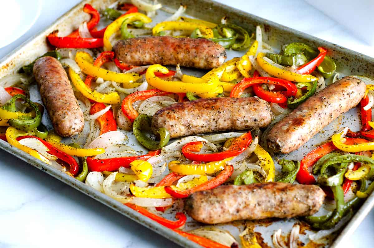 Metal sheet pan on a countertop with roasted sausages, red green and yellow peppers, and onions