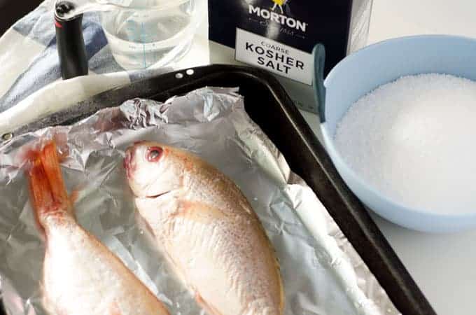 Pan lined with foil with two red snappers on it, with water, salt and a bowl in the background