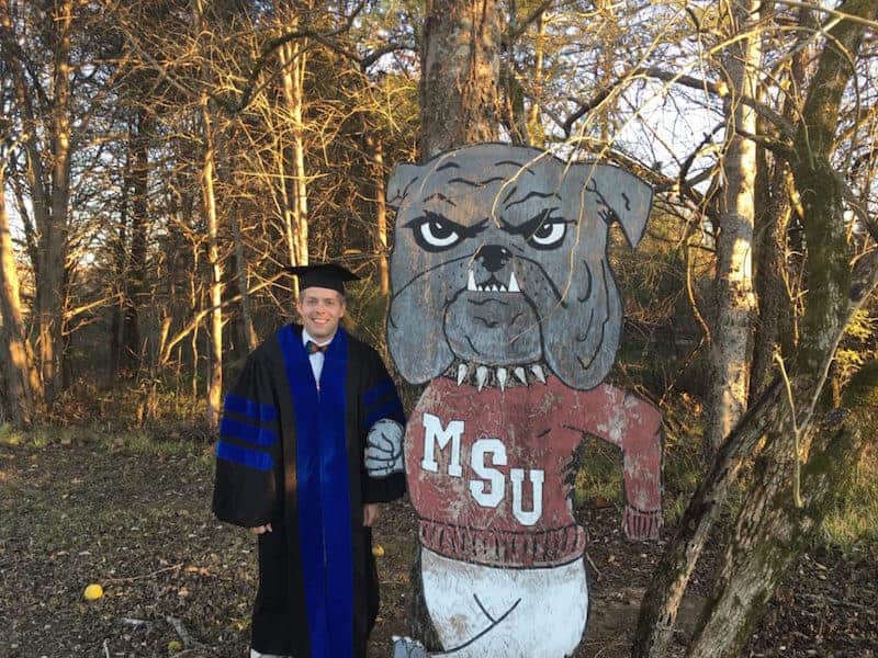 Man in graduation gown standing with MSU bulldog cutout in woods