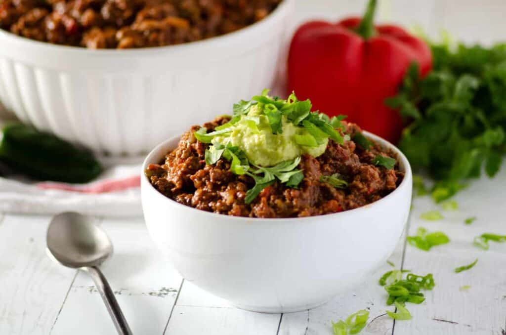 Side view of white bowl of paleo chili topped with an avocado cream; large serving bowl of chili in the background with red pepper and bunch of cilantro