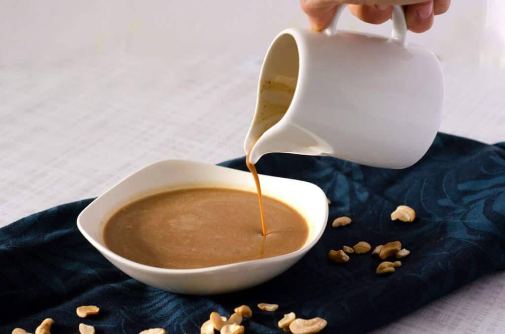 hand holding a white pitcher pouring pad thai peanut sauce into a bowl