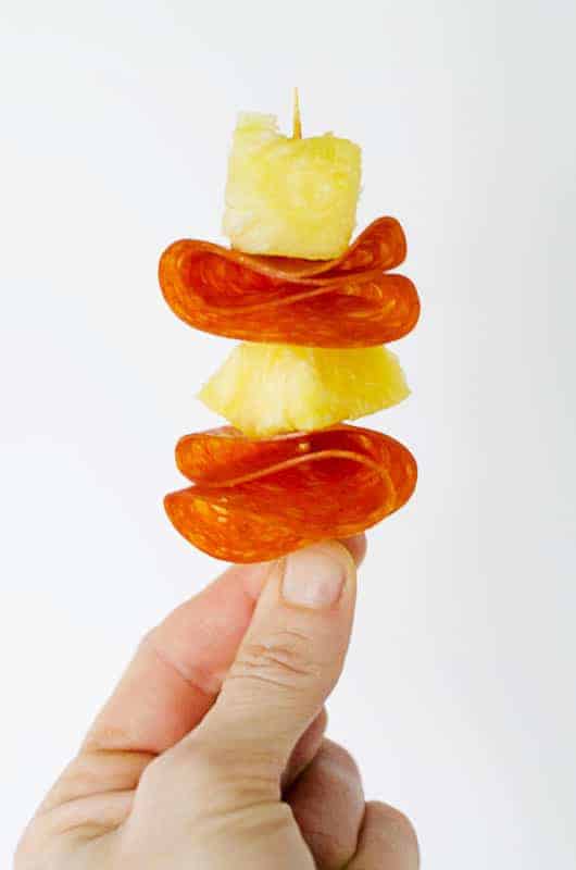 close up of a hand pinching a toothpick with pepperoni and pineapple skewered on it