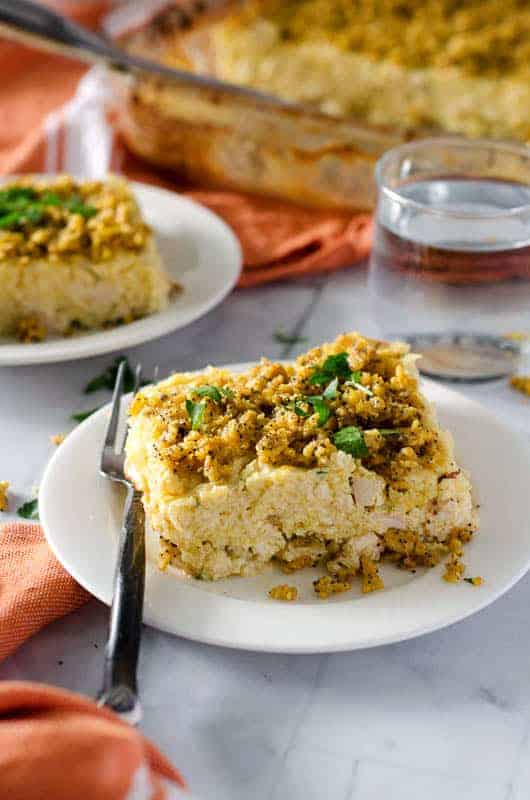 Side view of a slice of healthy chicken poppyseed casserole on a plate with a fork with a glass of water in the background