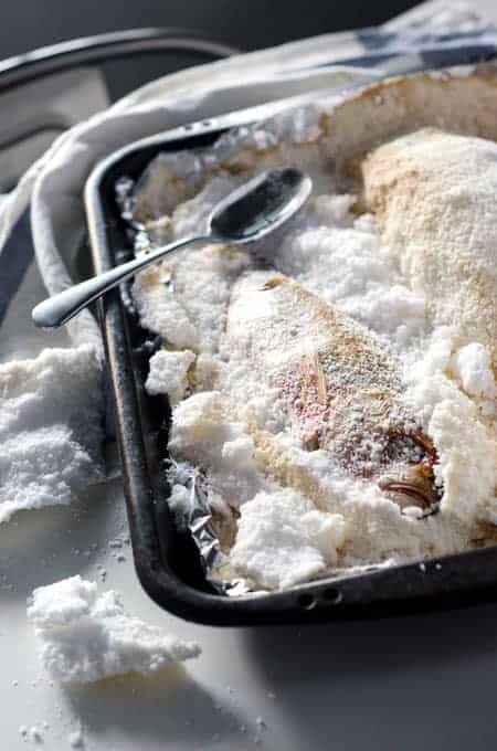 Red snapper baked in a salt crust on a baking sheet, with the salt crust broken open and chunks of salt surrounding