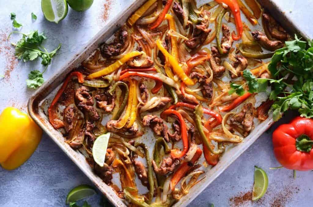 overhead view of a sheet pan covered in coked chicken, bell peppers and onions with slices of lime