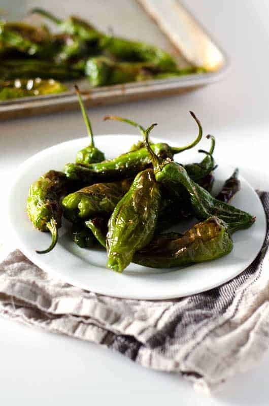 Side view of roasted shishito peppers on a plate with pan in the background