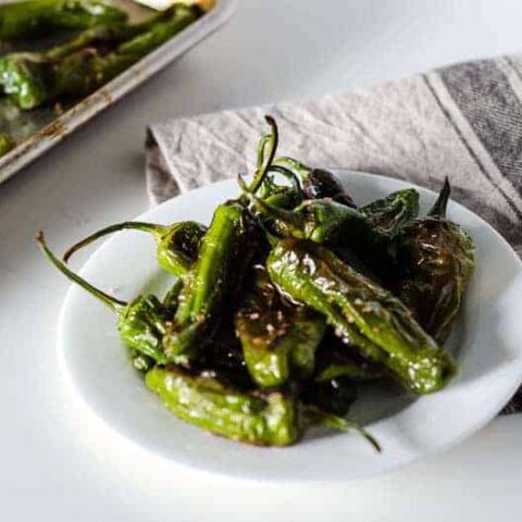 White plate of roasted shishito peppers with pan in the background