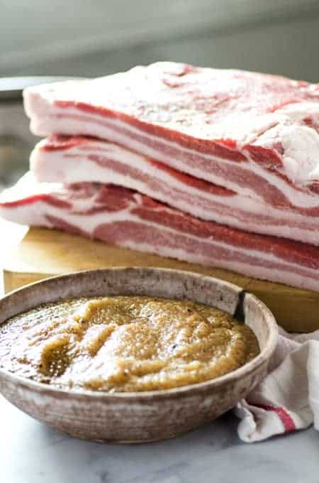 bowl of pork belly cure with slabs of pork belly on cutting board behind it