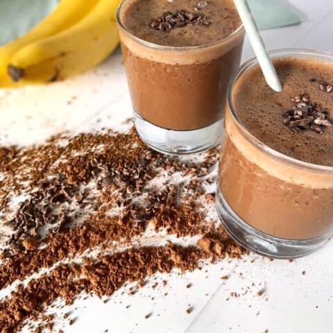 two banana coffee smoothies on countertop surrounded by cocoa powder, coffee, and bananas