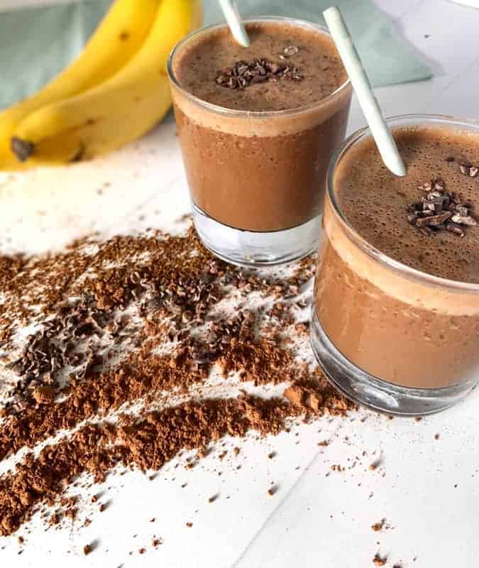 two banana coffee smoothies on countertop surrounded by cocoa powder, coffee, and bananas