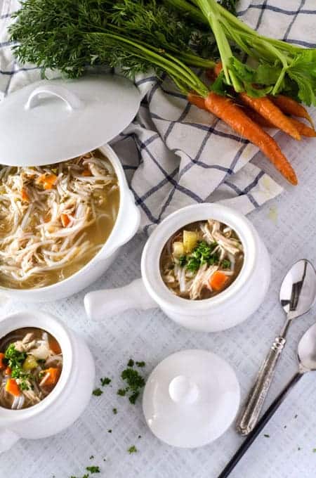 Overhead view of 2 bowls of paleo chicken noodle soup with a serving bowl of soup next to them surrounded by spoons, a dish towel and fresh carrots and celery