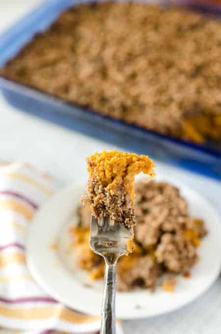 fork being held vertically with a bite of sweet potato casserole with pecan topping on it, with plate and casserole dish in the background