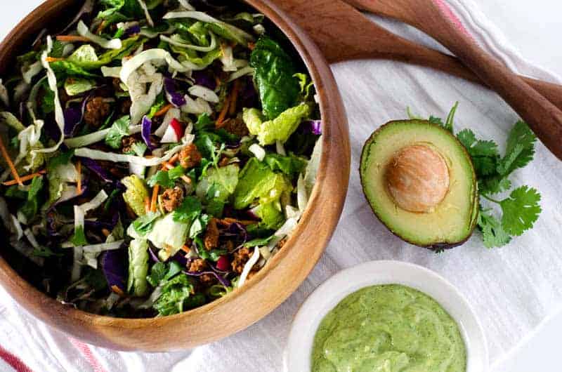 overhead view of wooden salad bowl filled with Whole30 taco salad surrounded by salad tongs, avocado, cilantro, and a bowl of dressing