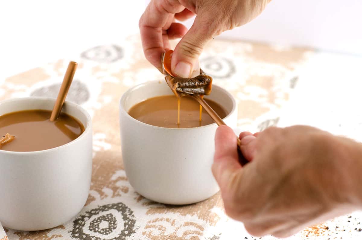 Two white mugs with dirty chai latte in them with a woman's hands squeezing out a chai tea bag over one.