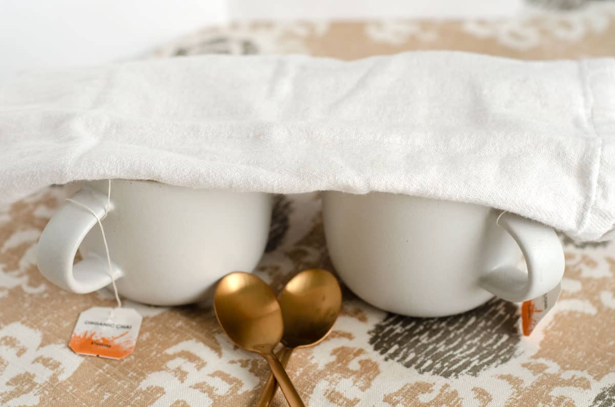 Side view of two white mugs with two gold spoons in front and a white tea towel folded up over the mugs