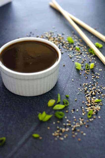 bowl of teriyaki sauce surrounded by sesame seeds, green onions and chopsticks