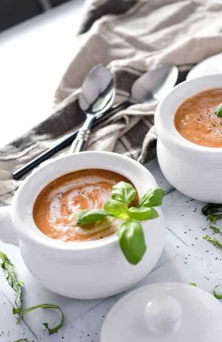 two white crocks filled with tomato basil soup with spoons and a napkin in the background