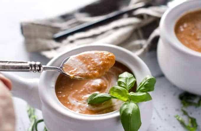 white crock of tomato basil soup with a hand holding a spoonful of soup above the dish