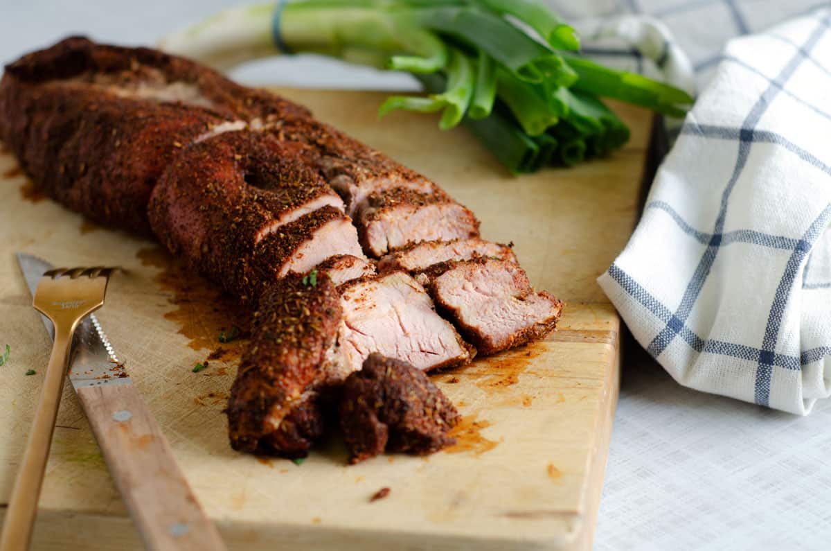 smoked pork tenderloin sliced on cutting board with green onions and plaid dish towel in background