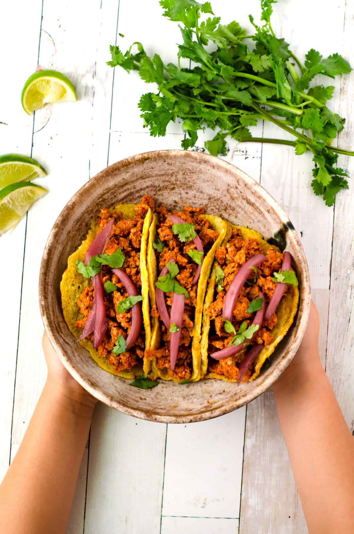 Three chorizo breakfast tacos topped with pickled red onions in a bowl held by a child's hands, surrounded with lime slices and cilantro