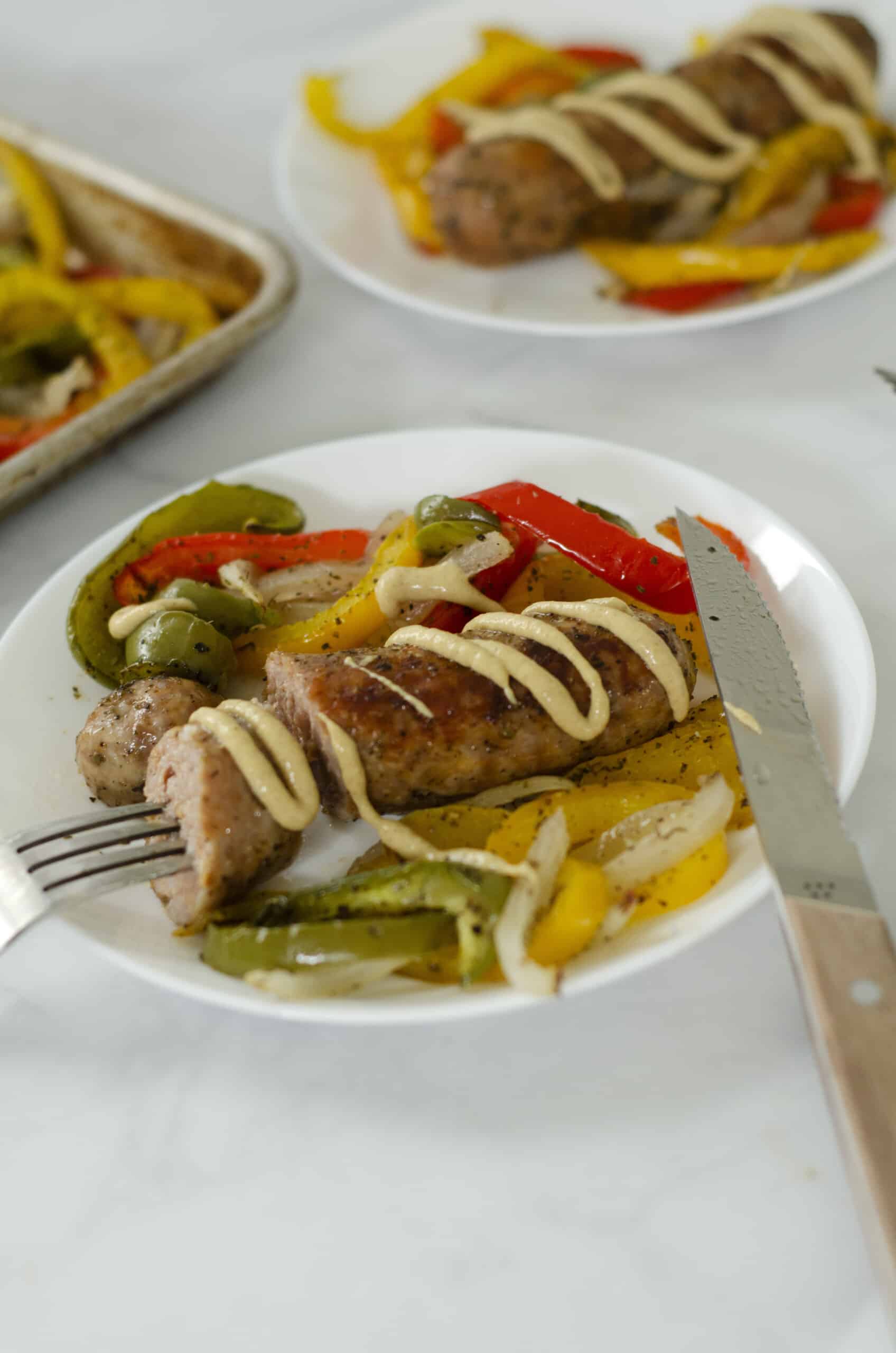 Close up of sausage peppers and onions on a plate with a drizzle of dijon mustard and a knife and fork